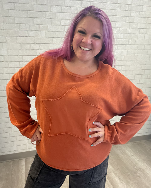 Person wearing an orange sweatshirt with a star design against a light brick wall.