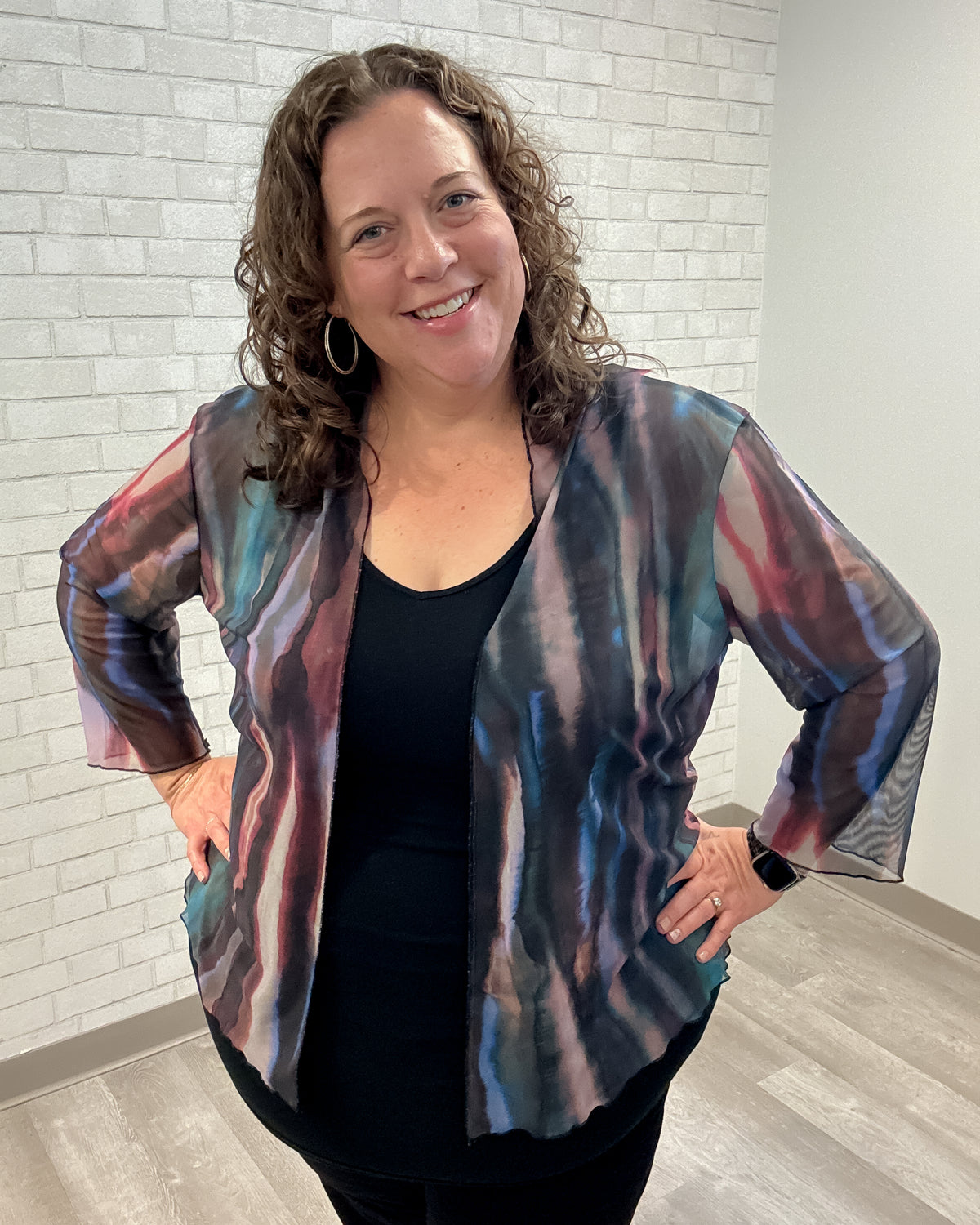 Woman wearing a colorful jacket over a black top against a white brick wall.