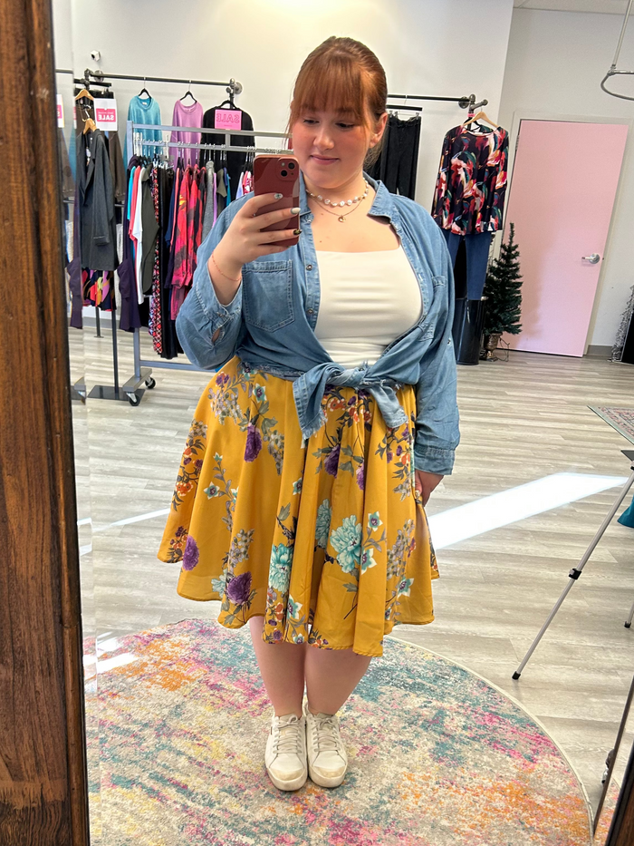 Person taking a mirror selfie wearing a yellow floral skirt and blue denim jacket in a clothing store.
