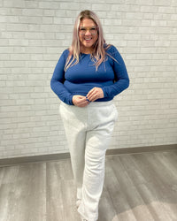 Person wearing a blue top and white pants standing against a light brick wall.