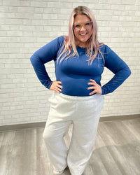 Person wearing a blue long-sleeve top and white sweatpants against a light brick wall.