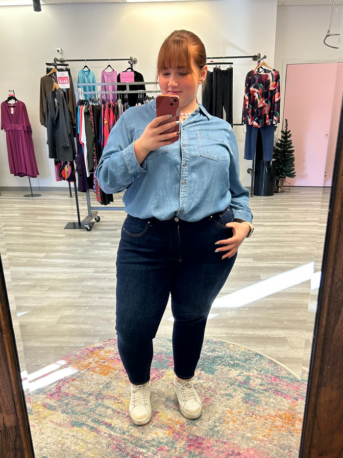 Person taking a mirror selfie wearing a blue denim shirt and dark jeans in a clothing store.